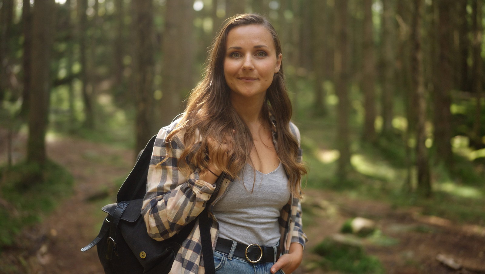 kobieta w wieku 30 plus w koszuli w kratę z plecakiem stoi w lesie i zalotnie się uśmiecha