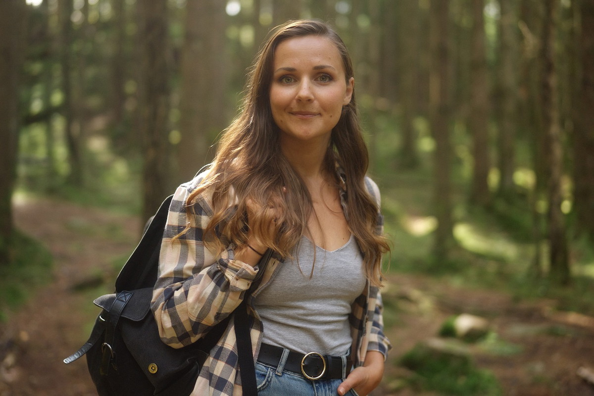 kobieta w wieku 30 plus w koszuli w kratę z plecakiem stoi w lesie i zalotnie się uśmiecha