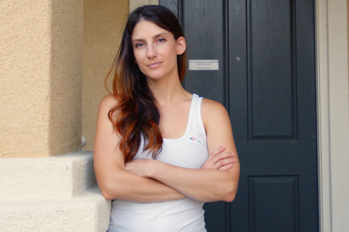 kobieta w wieku 30 plus z długimi włosami w białym ropie stoi na tle drzwi ze skrzyżowanym rękami i z poważną miną