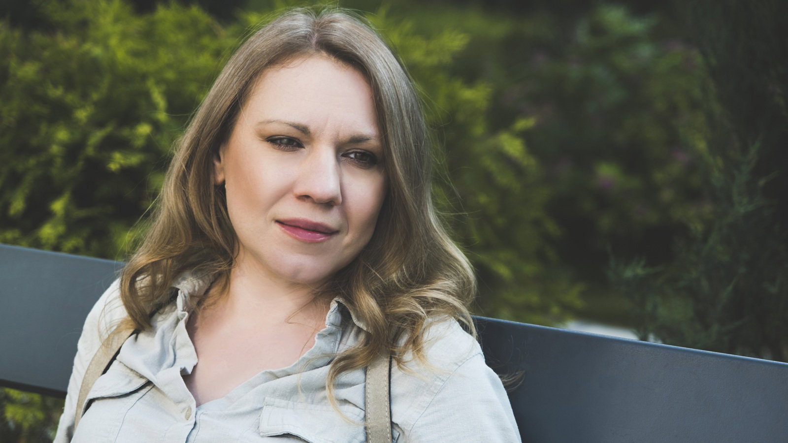 kobieta w wieku 40 plus siedzi na ławce na tle zieleni z plecakiem na plecach zamyślona