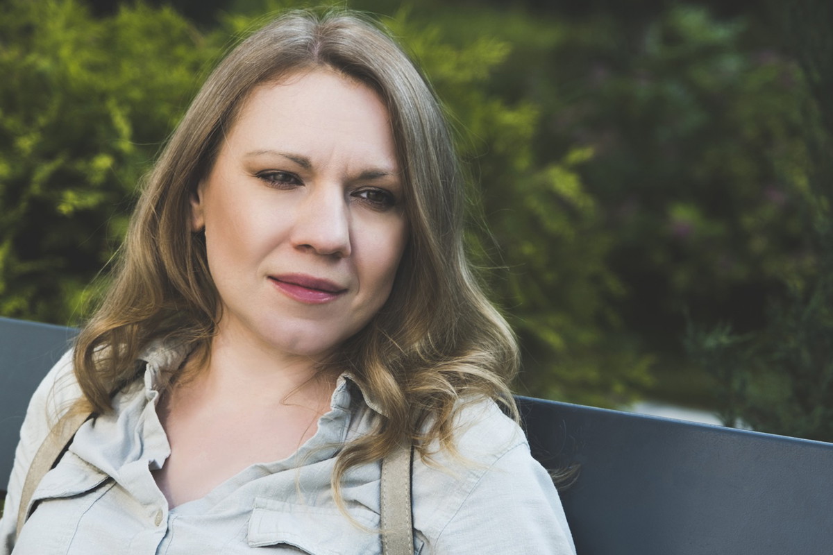 kobieta w wieku 40 plus siedzi na ławce na tle zieleni z plecakiem na plecach zamyślona