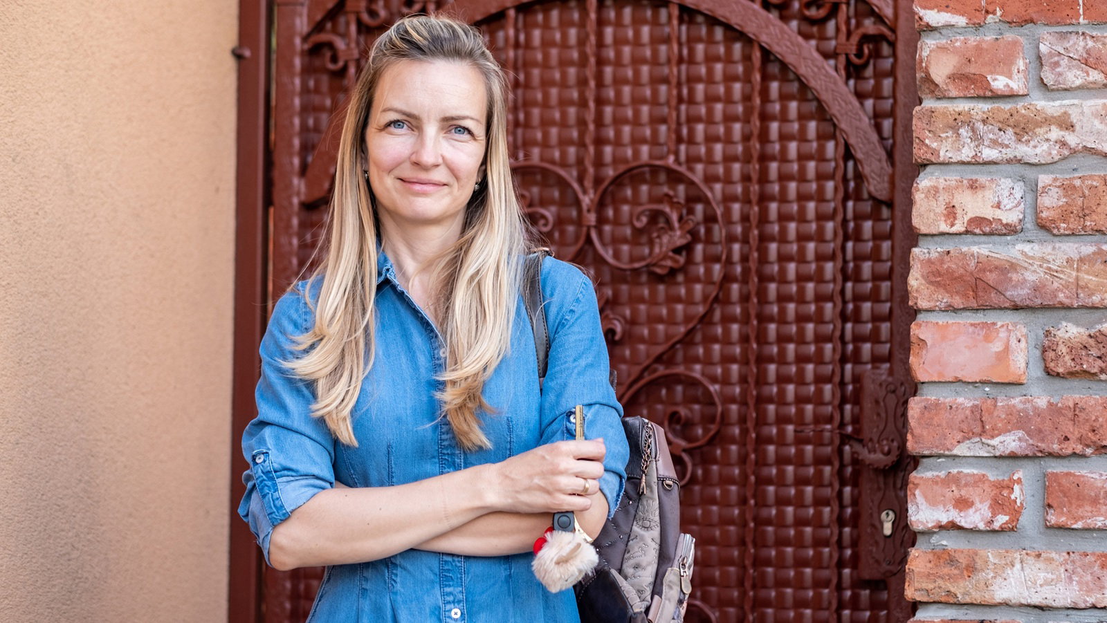 kobieta w wieku 40 plus w jeansowej sukience stoi na tle zdobionych drzwi z lekkim uśmiechem