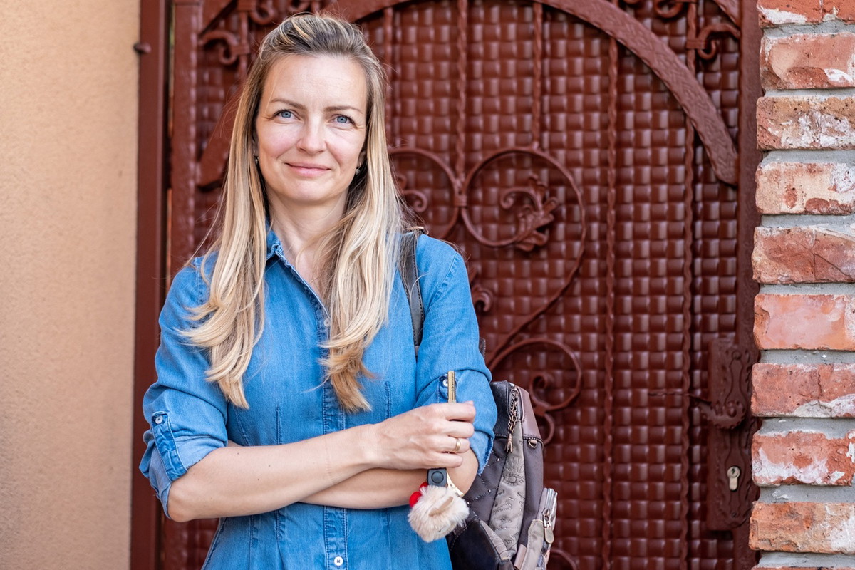 kobieta w wieku 40 plus w jeansowej sukience stoi na tle zdobionych drzwi z lekkim uśmiechem