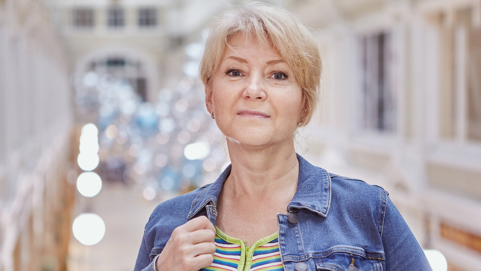 kobieta w wieku 50 plus blondynka stoi na tle rozświetlonego budynku z lekko rozmarzonym uśmiechem