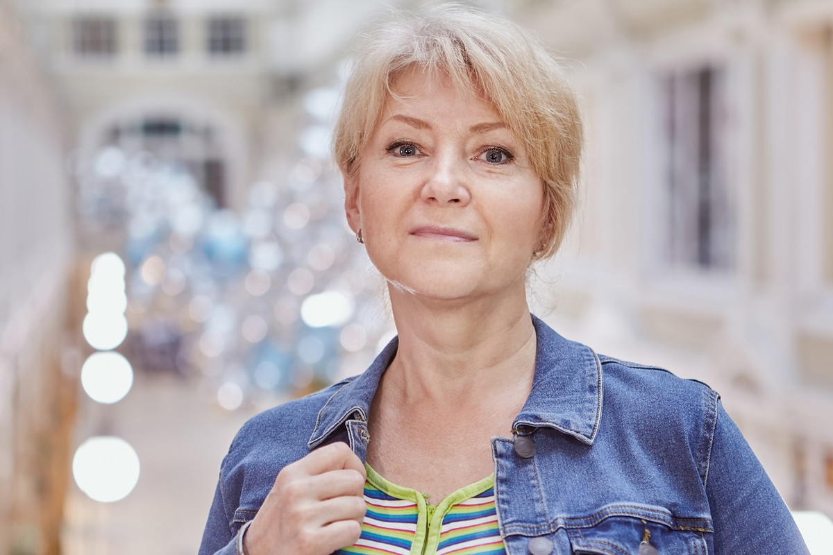 kobieta w wieku 50 plus blondynka stoi na tle rozświetlonego budynku z lekko rozmarzonym uśmiechem