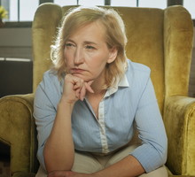 kobieta w wieku 50 plus blondynka w niebieskiej koszuli siedzi na welurowym fotelu zamyślona i zdenerwowana
