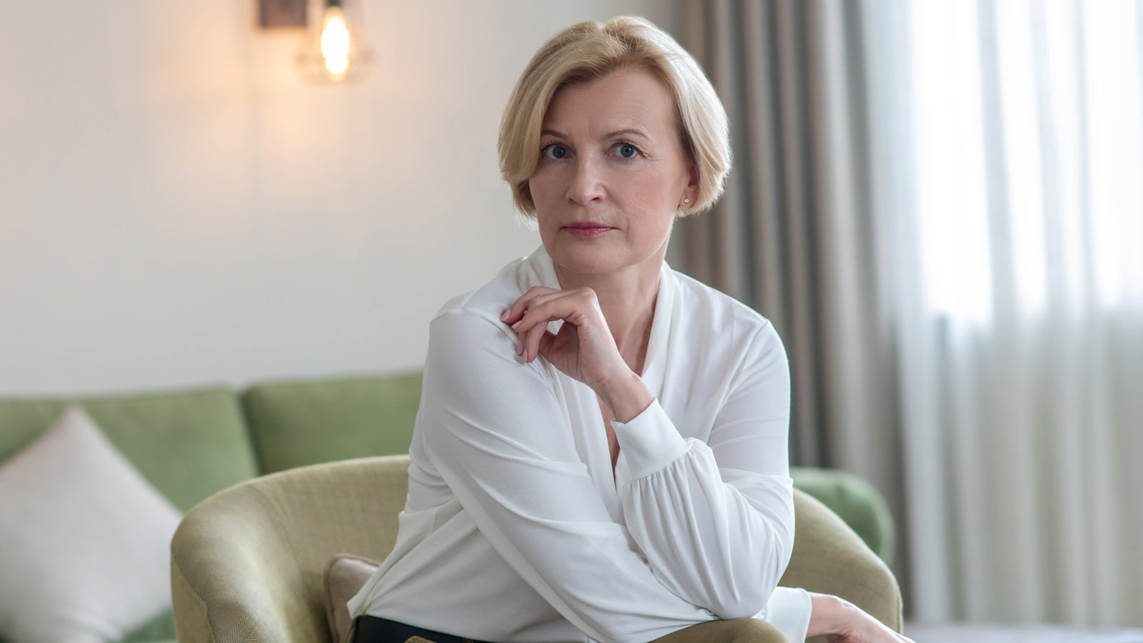 kobieta w wieku 55 plus blondynka w białej bluzce siedzi w pokoju z poważną nieco wystraszoną miną