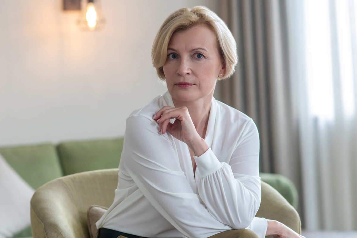 kobieta w wieku 55 plus blondynka w białej bluzce siedzi w pokoju z poważną nieco wystraszoną miną