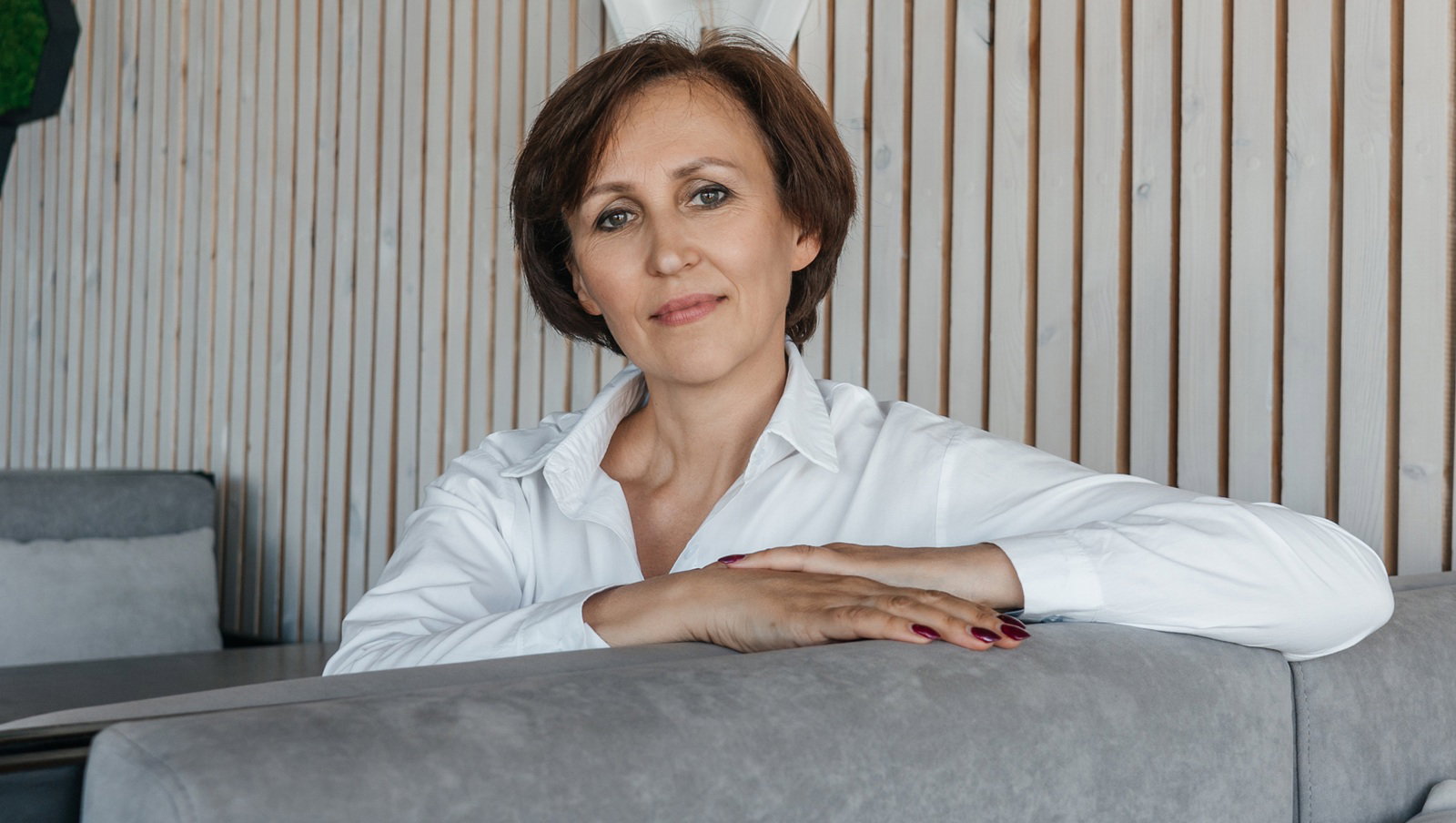 kobieta w wieku 55 plus w białej koszuli siedzi w na sali na kanapie przy stoliku zamyślona z lekkim uśmiechem