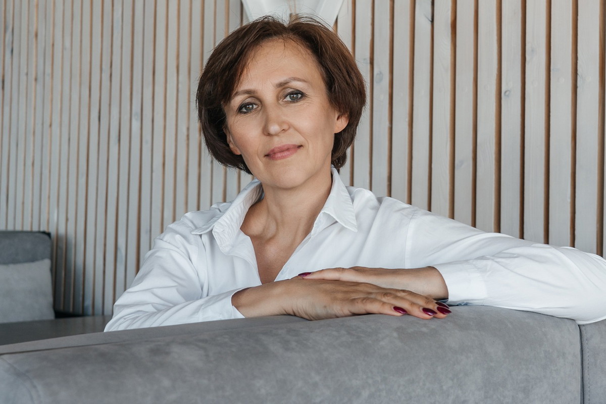 kobieta w wieku 55 plus w białej koszuli siedzi w na sali na kanapie przy stoliku zamyślona z lekkim uśmiechem