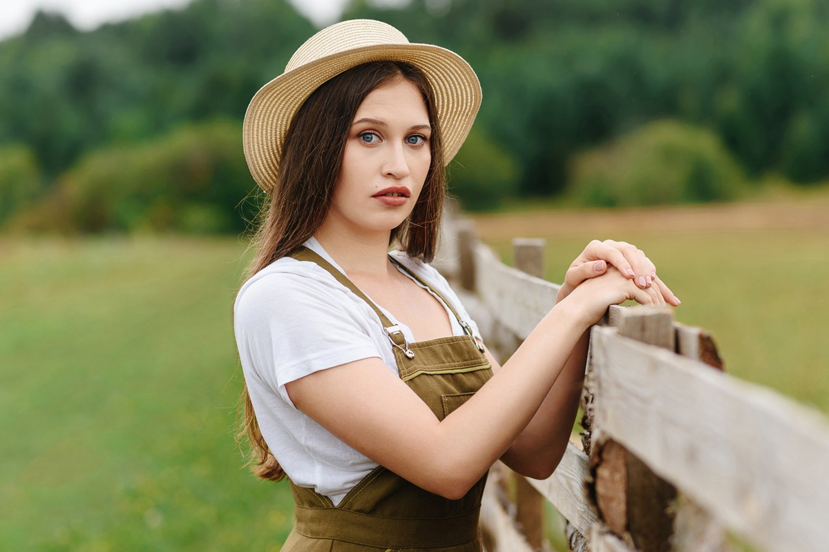 młoda kobieta w kapeluszu w wieku 25 plus stoi na tle wiejskiego pejzażu ze zdziwioną i zniesmaczoną miną