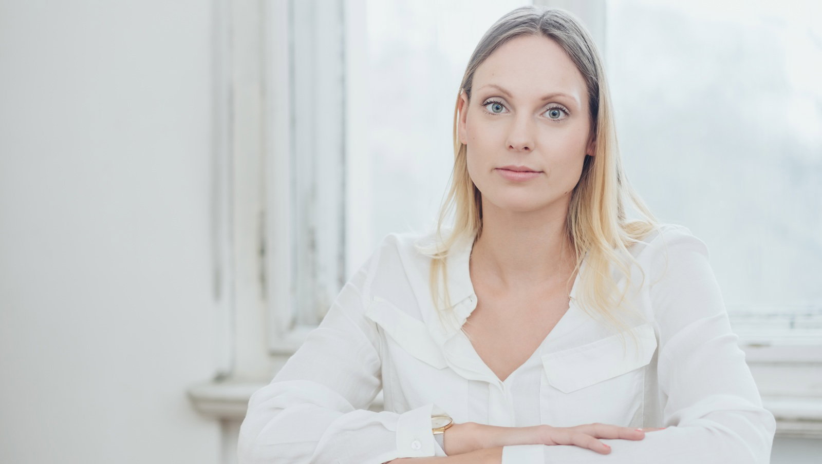 młoda kobieta w wieku 30 plus blondynka w białej koszuli siedzi na krześle na tle okna zamyślona