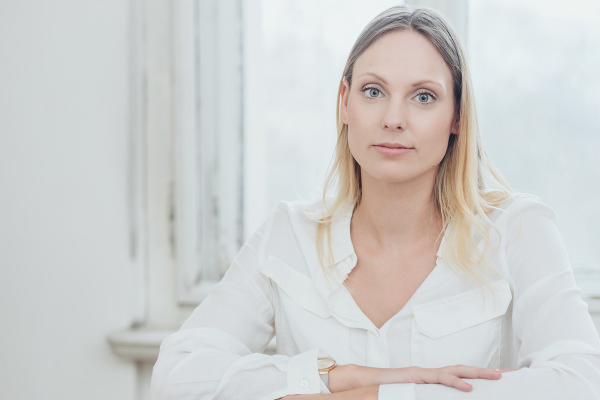 młoda kobieta w wieku 30 plus blondynka w białej koszuli siedzi na krześle na tle okna zamyślona