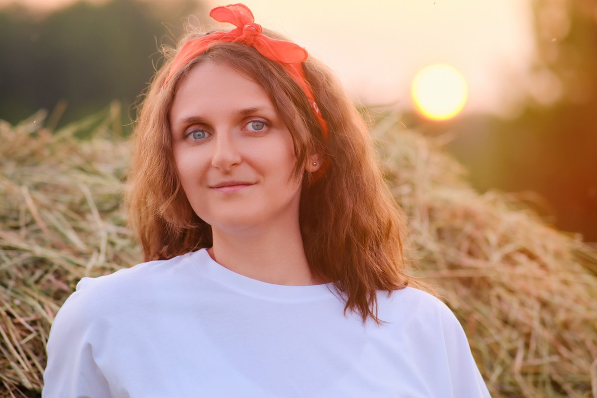 kobieta w wieku 35 plus z chusteczką na głowie i w białej bluzce stoi na tle zachodzącego słońca i siana