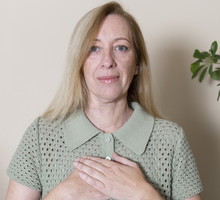 kobieta w wieku 50 plus z blond włosami w zielonej bluzce stoi z rękami na sercu i pogodną miną