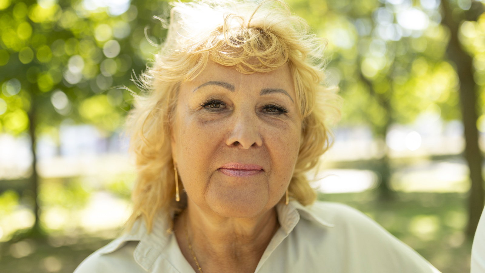 kobieta w wieku 60 plus blond włosy stoi na tle zieleni na zewnątrz z lekkim uśmiechem na twarzy