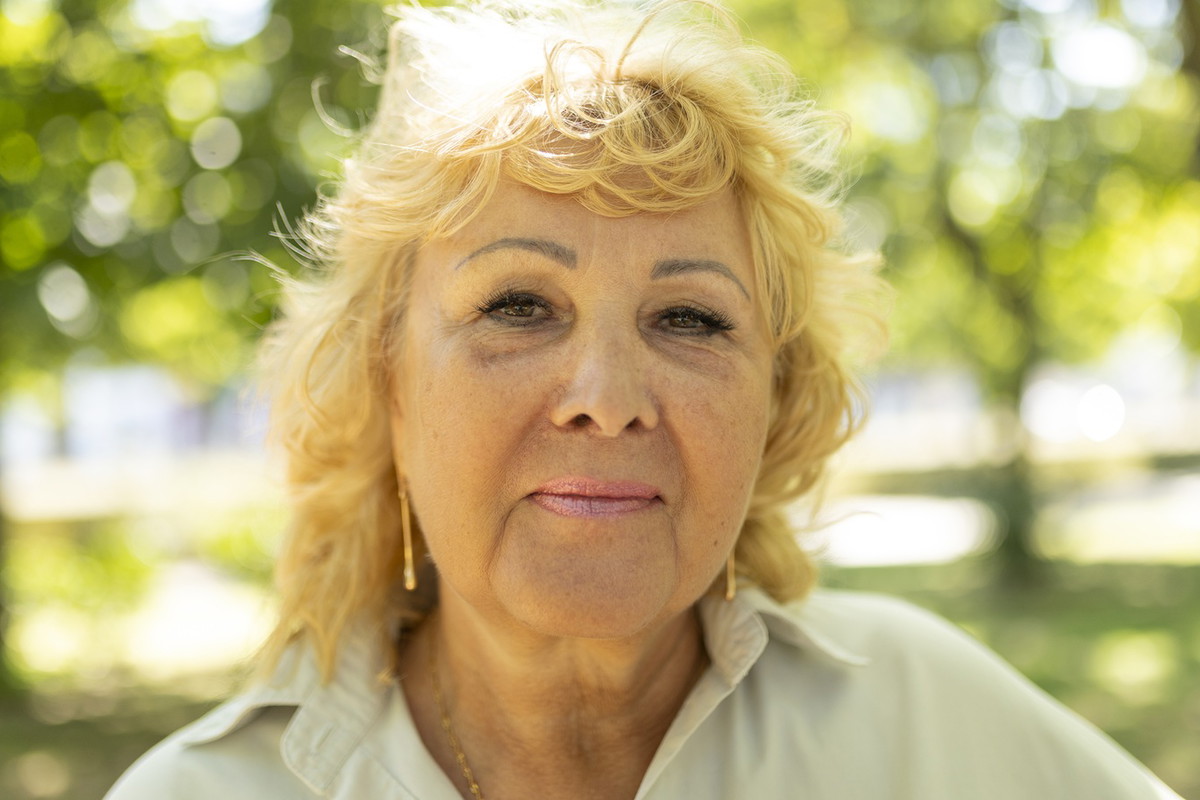 kobieta w wieku 60 plus blond włosy stoi na tle zieleni na zewnątrz z lekkim uśmiechem na twarzy