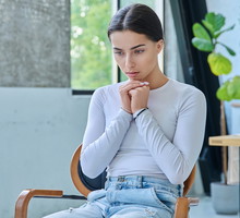 młoda dziewczyna w wieku około 19 plus w jeansach i koszulce longsleeve siedzi na krześle zamyślona i smutna