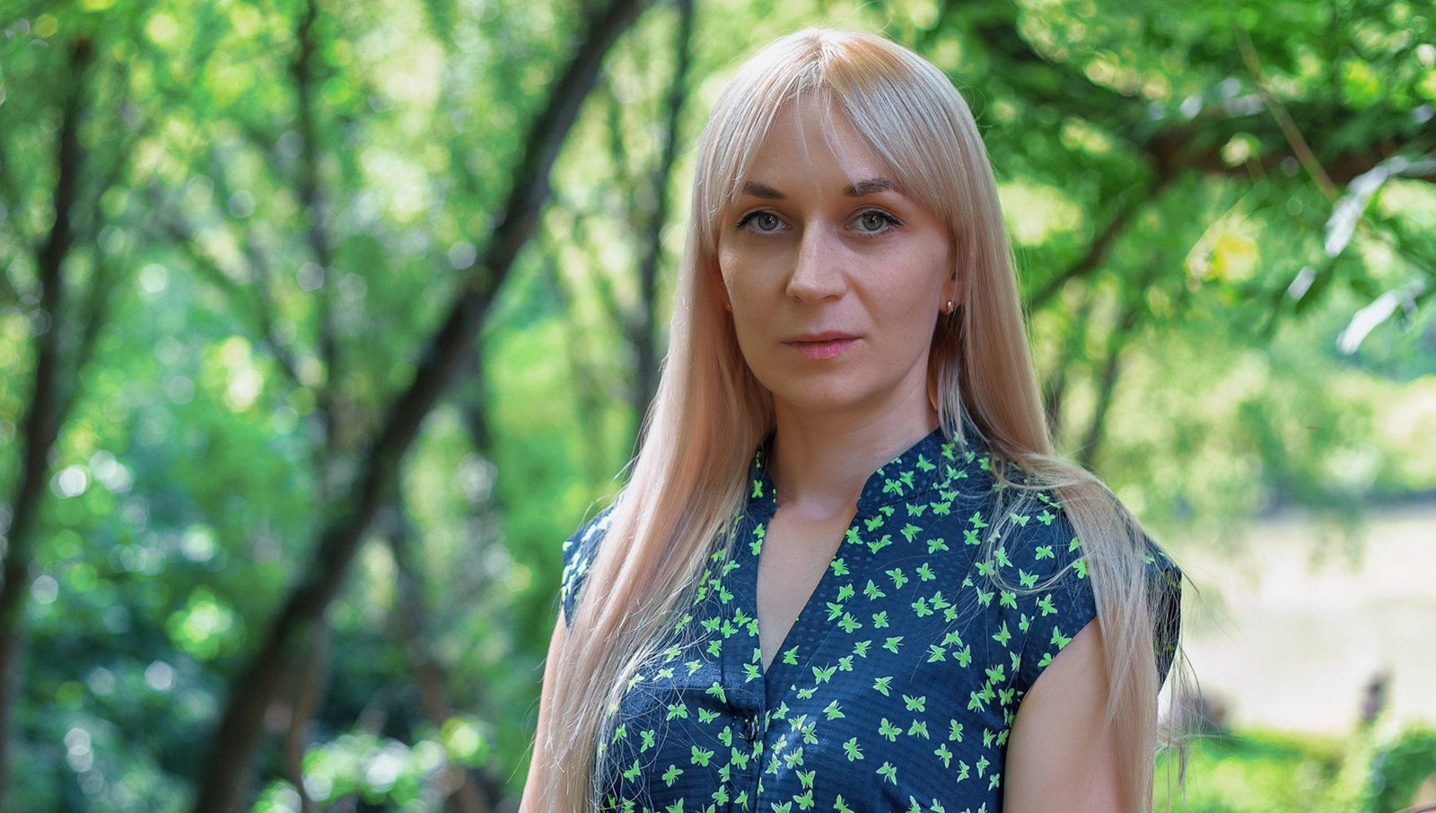 kobieta w wieku 25 plus blondynka w sukience w drobny wzorek stoi na tle zieleni z poważną miną