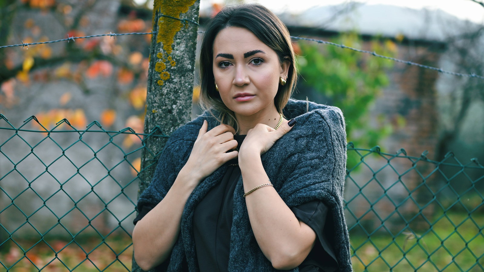kobieta w wieku 30 plus stoi przy płocie na tle ogordu zamyślona i smutna ze swetrem na ramionach