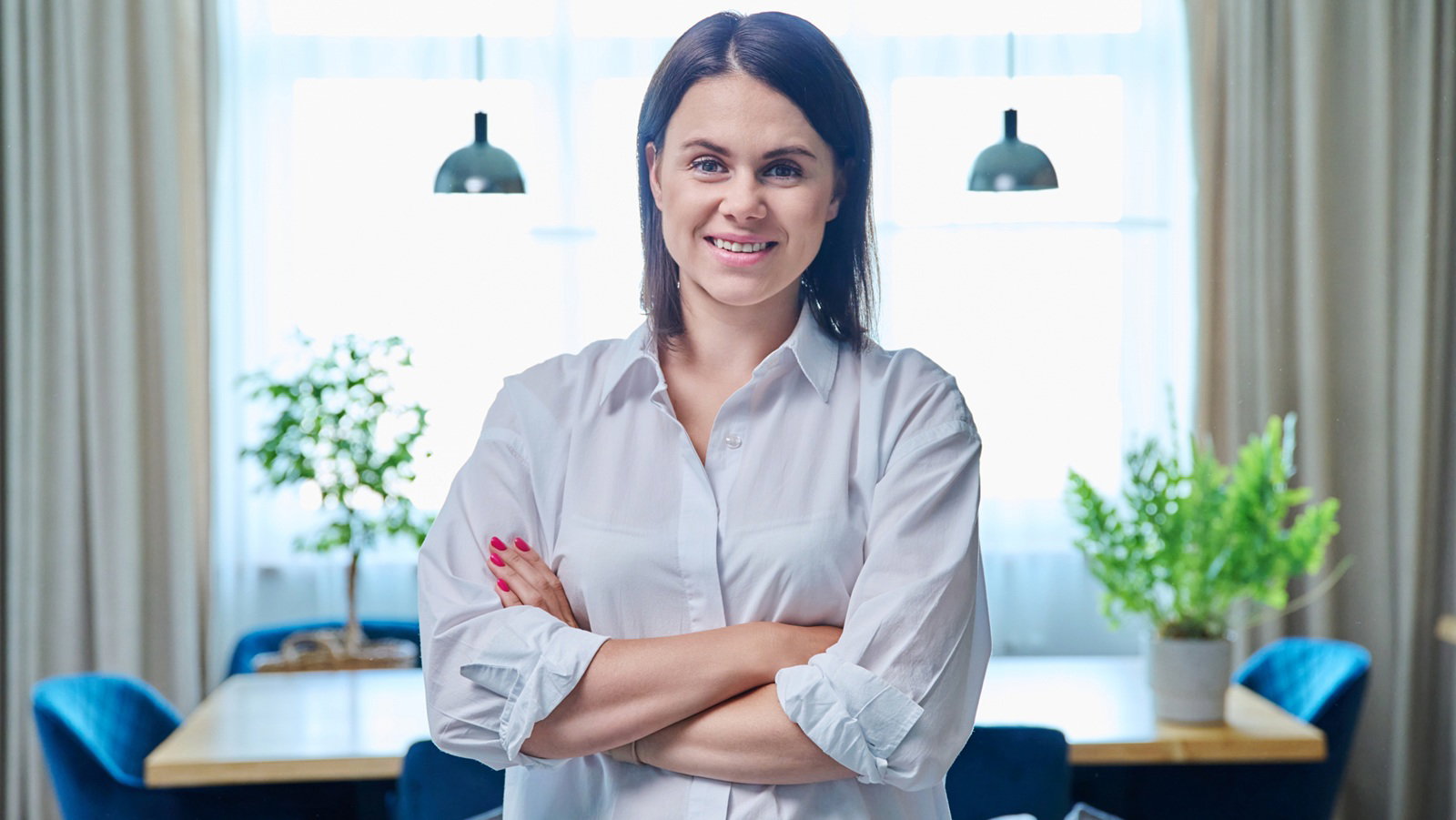 kobieta w wieku 30 plus w jasnej koszuli stoi na tle salonu zadowolona i z kpiącym uśmiechem