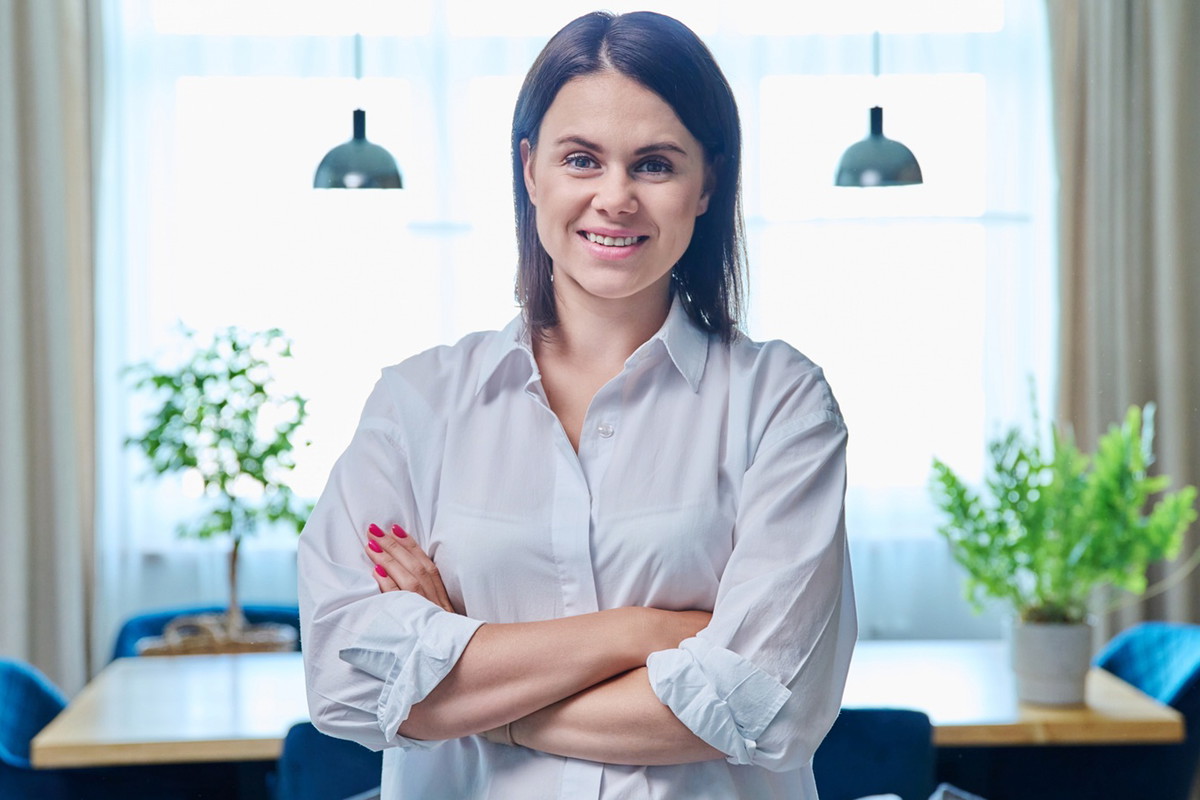 kobieta w wieku 30 plus w jasnej koszuli stoi na tle salonu zadowolona i z kpiącym uśmiechem