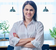 kobieta w wieku 30 plus w jasnej koszuli stoi na tle salonu zadowolona i z kpiącym uśmiechem