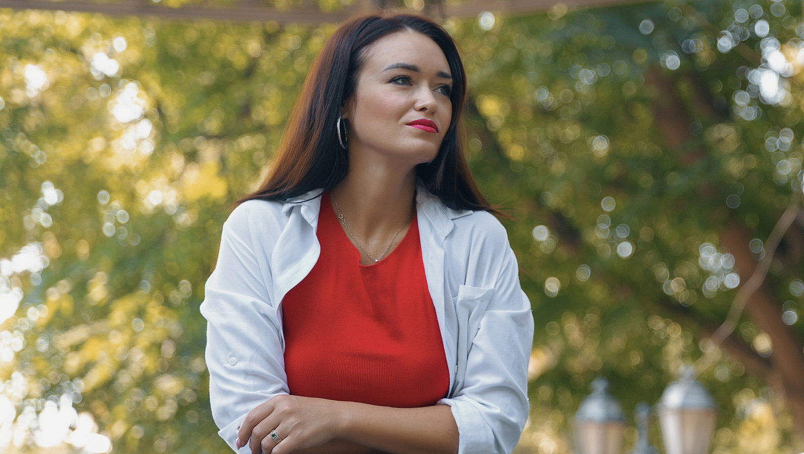kobieta w wieku 30 plus z ciemnymi włosami i w czerwonej bluzce stoi przy barierce na tle zieleni zamyślona