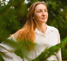 kobieta w wieku 30 plus z długimi włosami stoi za sosnami i patrzy w dal lekko się uśmiechając
