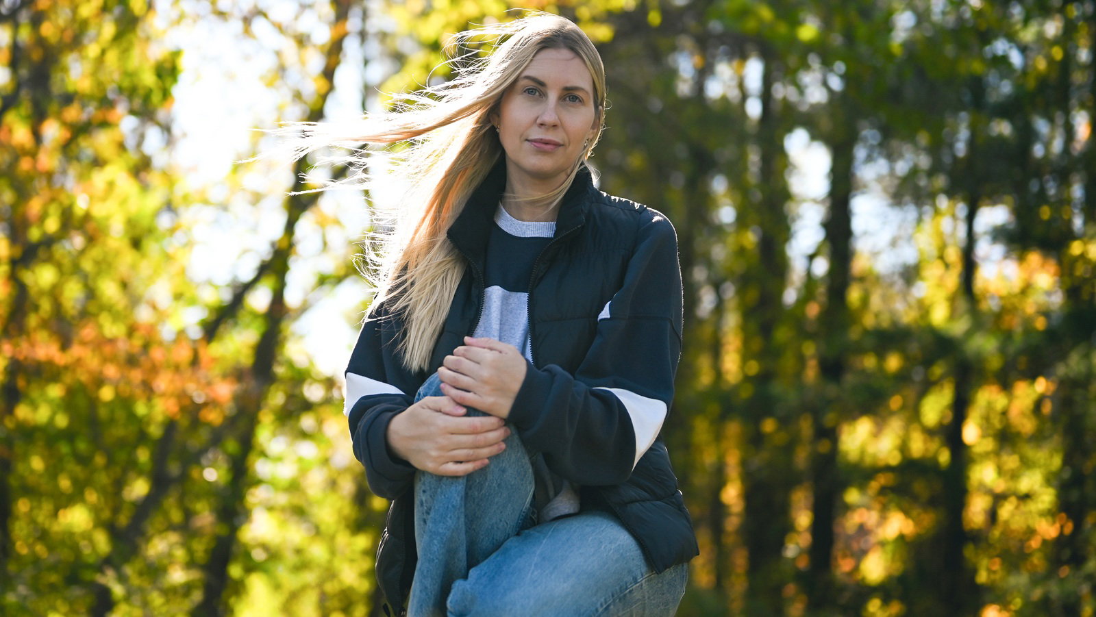 kobieta w wieku 30 plus z jasnymi długimi włosami kuca na tle lasu z poważną miną