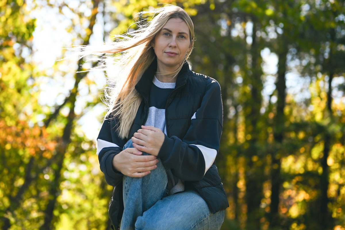 kobieta w wieku 30 plus z jasnymi długimi włosami kuca na tle lasu z poważną miną