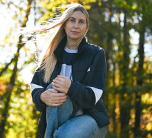 kobieta w wieku 30 plus z jasnymi długimi włosami kuca na tle lasu z poważną miną