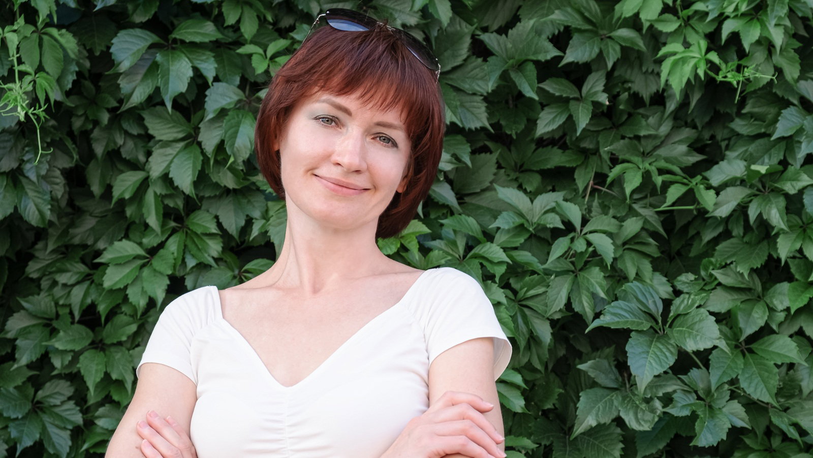 kobieta w wieku 40 plus w białej bluzce stoi na tle zielonych roścli i uśmiecha się lekko rozmarzona