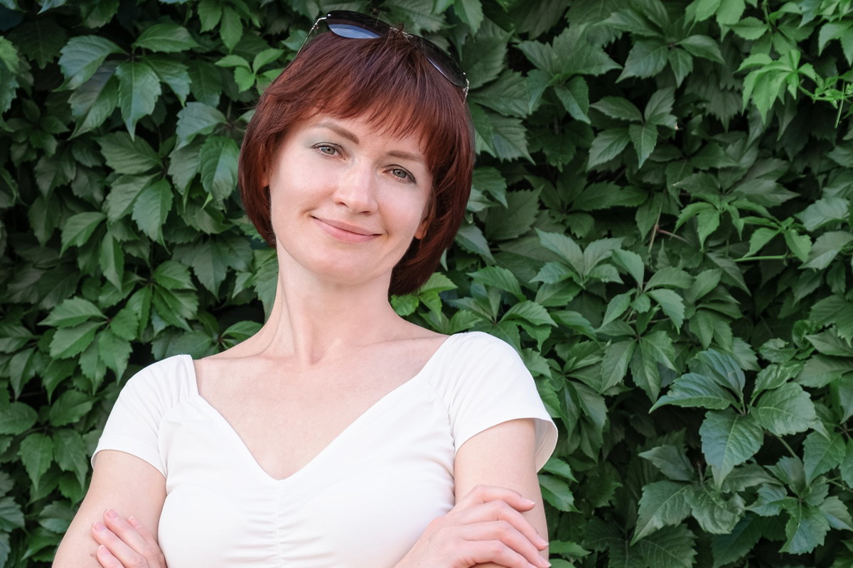 kobieta w wieku 40 plus w białej bluzce stoi na tle zielonych roścli i uśmiecha się lekko rozmarzona