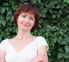 kobieta w wieku 40 plus w białej bluzce stoi na tle zielonych roścli i uśmiecha się lekko rozmarzona