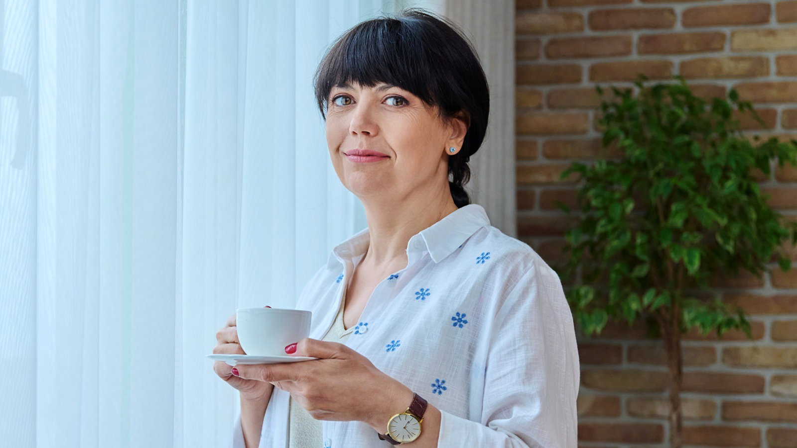 kobieta w wieku 50 plus stoi przy onie z filiżanką kawy z lekkim uśmiechem zamyślona
