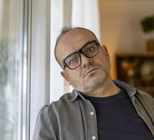 mężczyzna w wieku 50 plus w okularach i rozpiętej koszuli ze zmartwioną poważną miną stoi przy oknie