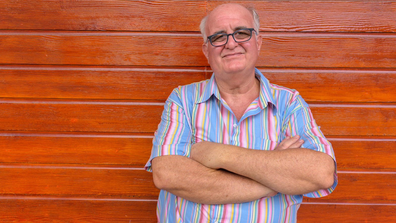 mężczyzna w wieku 60 plus w okularach łysiejący stoi na tle drewnianej ściany ze zmieszaną miną