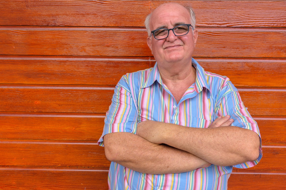 mężczyzna w wieku 60 plus w okularach łysiejący stoi na tle drewnianej ściany ze zmieszaną miną