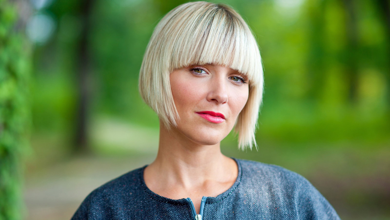 młoda kobieta w wieku 30 plus blondynka w fryzurze typu bob stoi na tle zieleni ze zrezygnowaną miną