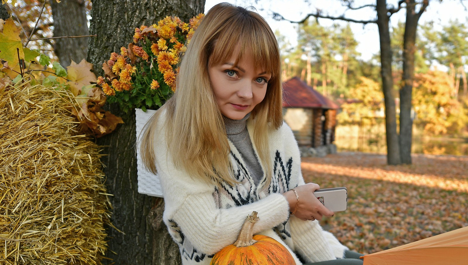 kobieta w wieku 30 plus blondynka siedzi pod drzewem z dynią przed sobą, siankiem i jesiennymi kwiatami ze sceptyczną miną