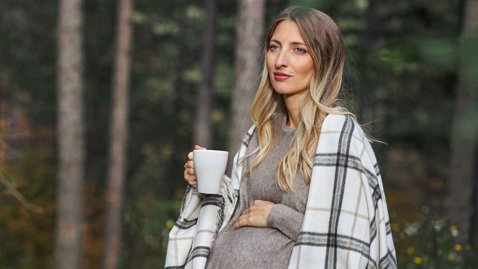 kobieta w wieku 30 plus blondynka w ciąży siedzi na tle lasu okryta kocykiem z kubkiem w ręku zamyślona