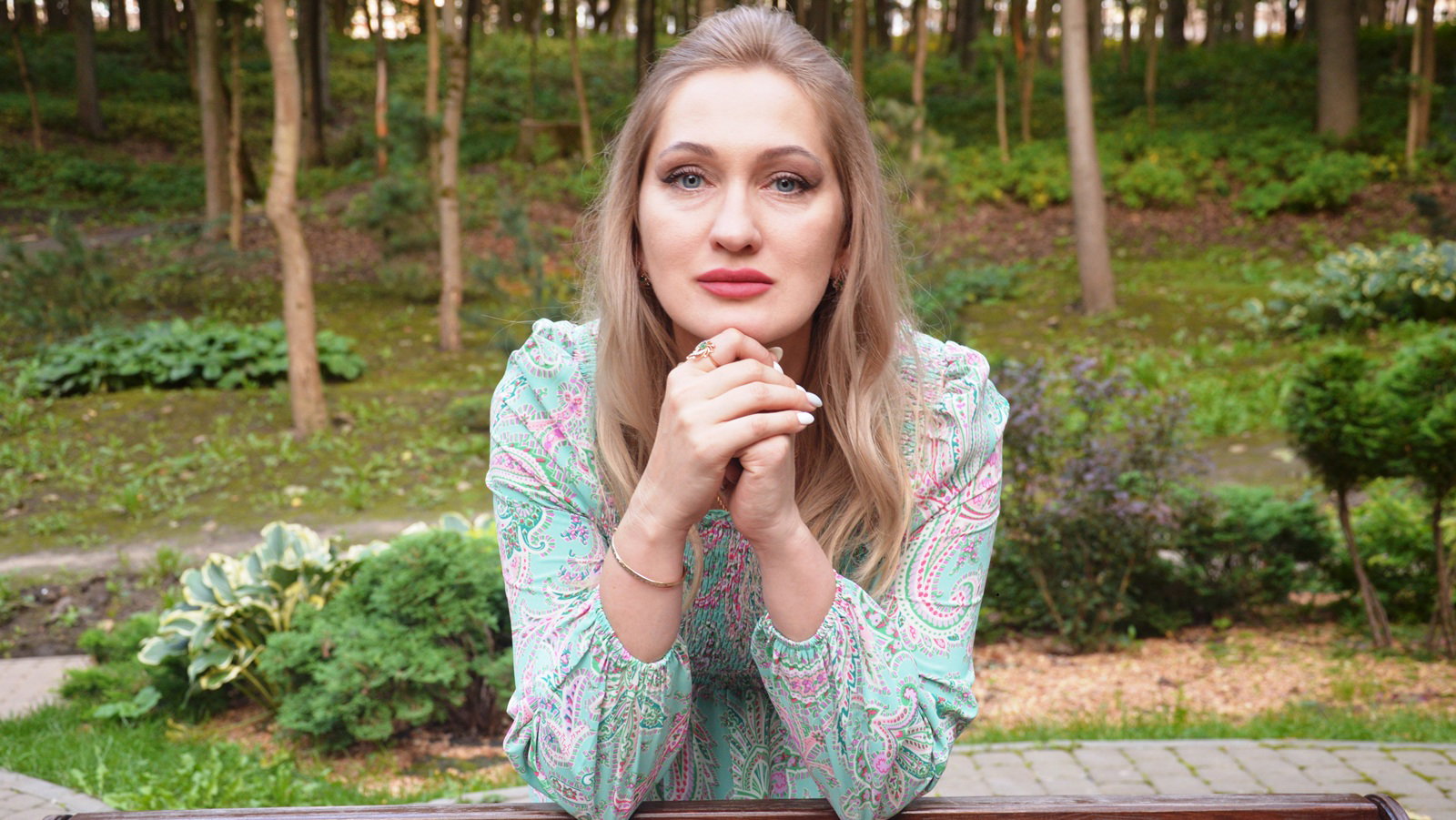 kobieta w wieku 30 plus blondynka we wzorzystj sukience siedzi na tle działki przy drewniany stole zamyślona
