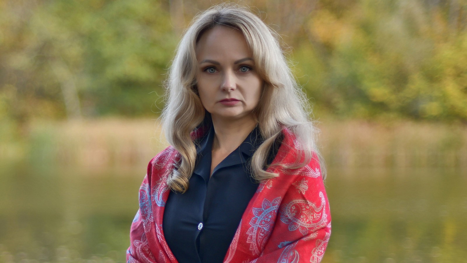 kobieta w wieku 40 plus blondynka w czarnej sukience i czerwonej wzorzystej chuście stoi na tle jesiennego pejzażu poważna i zamyślona
