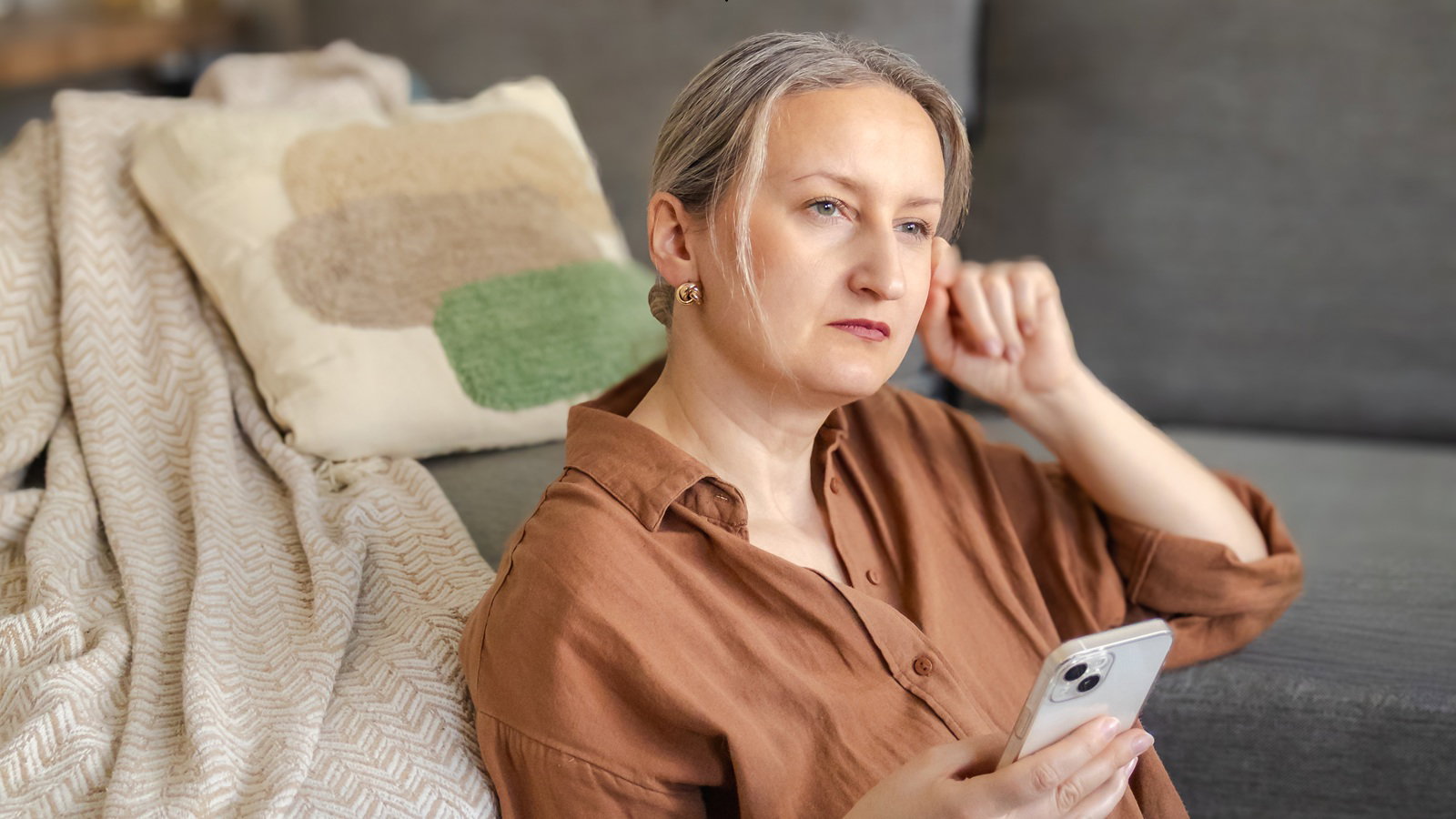 kobieta w wieku 40 plus w karmelowej koszuli siedzi na podłodze z telefonem w ręku zamyślona i smutna