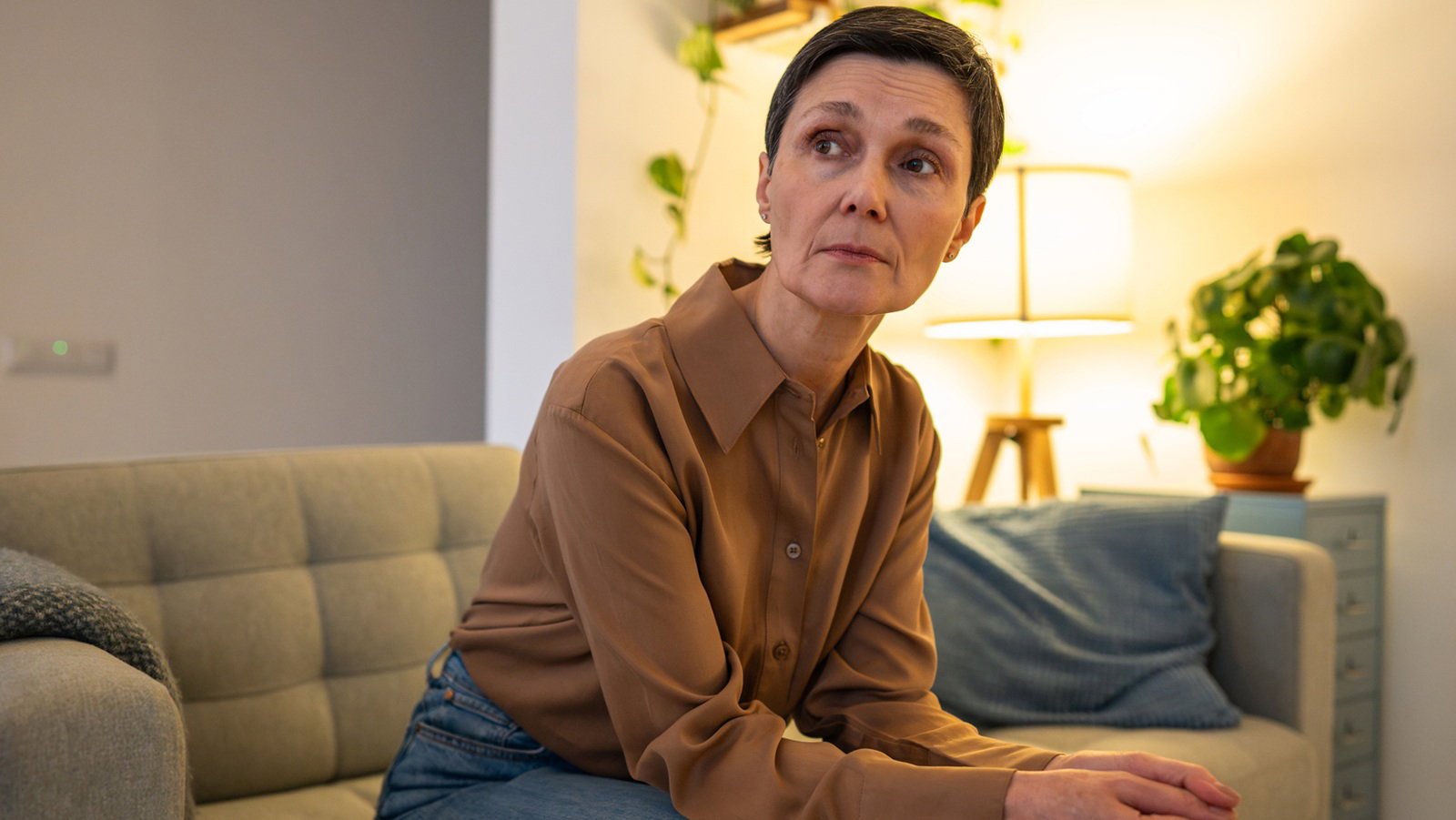 kobieta w wieku 50 plus w krótkiej fryzurze i karmelowej koszuli siedzi na kanapie załamana i smutna