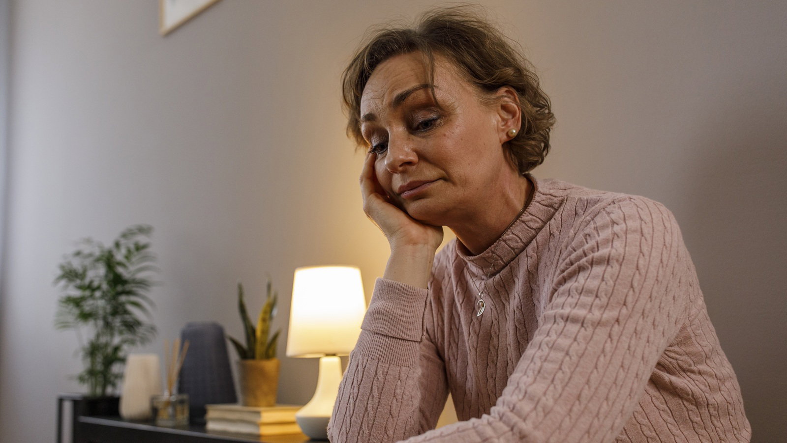 kobieta w wieku 50 plus we wrzosowym sweterku siedzi pod ścianą w salonie załamana i smutna