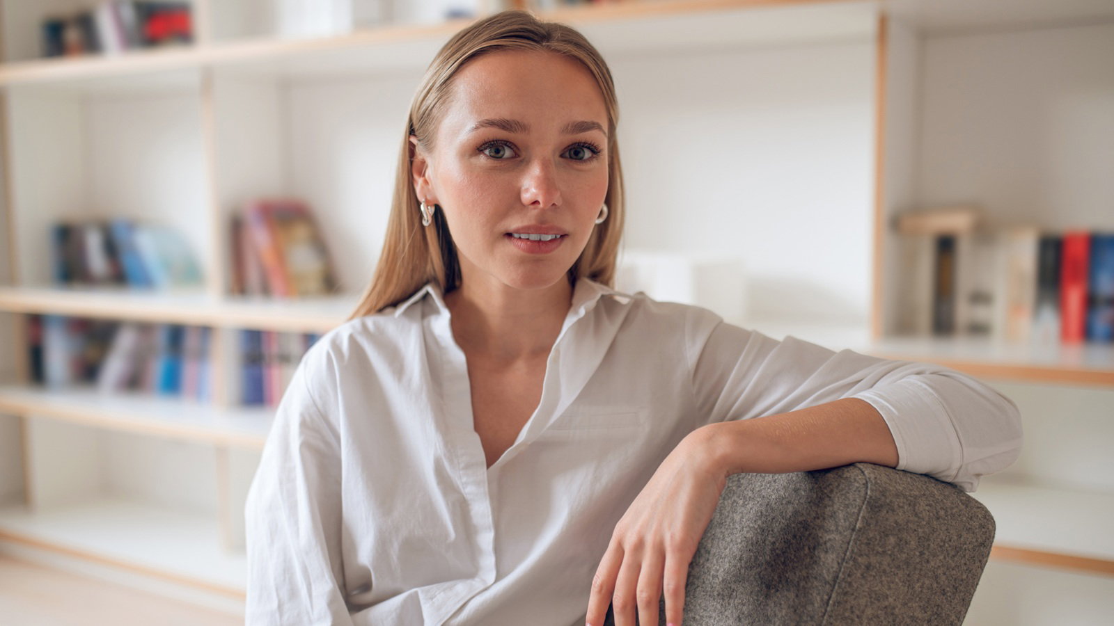 kobieta w wieku 25 plus blondynka z długimi włosami w białej koszuli siedzi na tle półek z książkami z niepewna miną