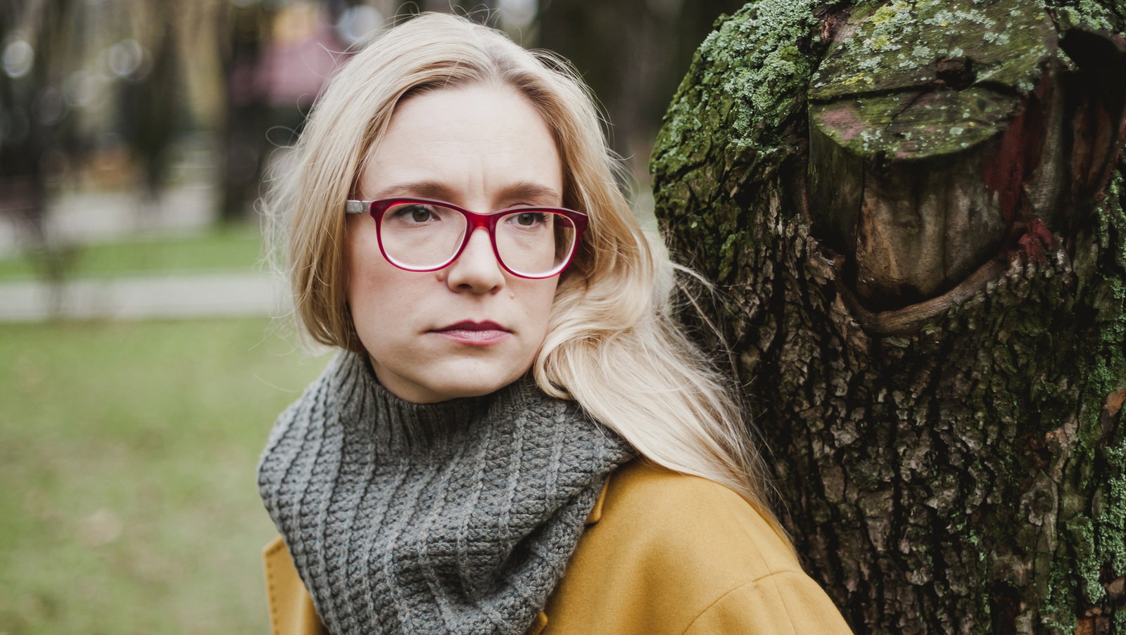 kobieta w wieku 30 plus blondynka w okularach i żółtym płaszczu stoi przy drzewie zamyślona smutna i poważna