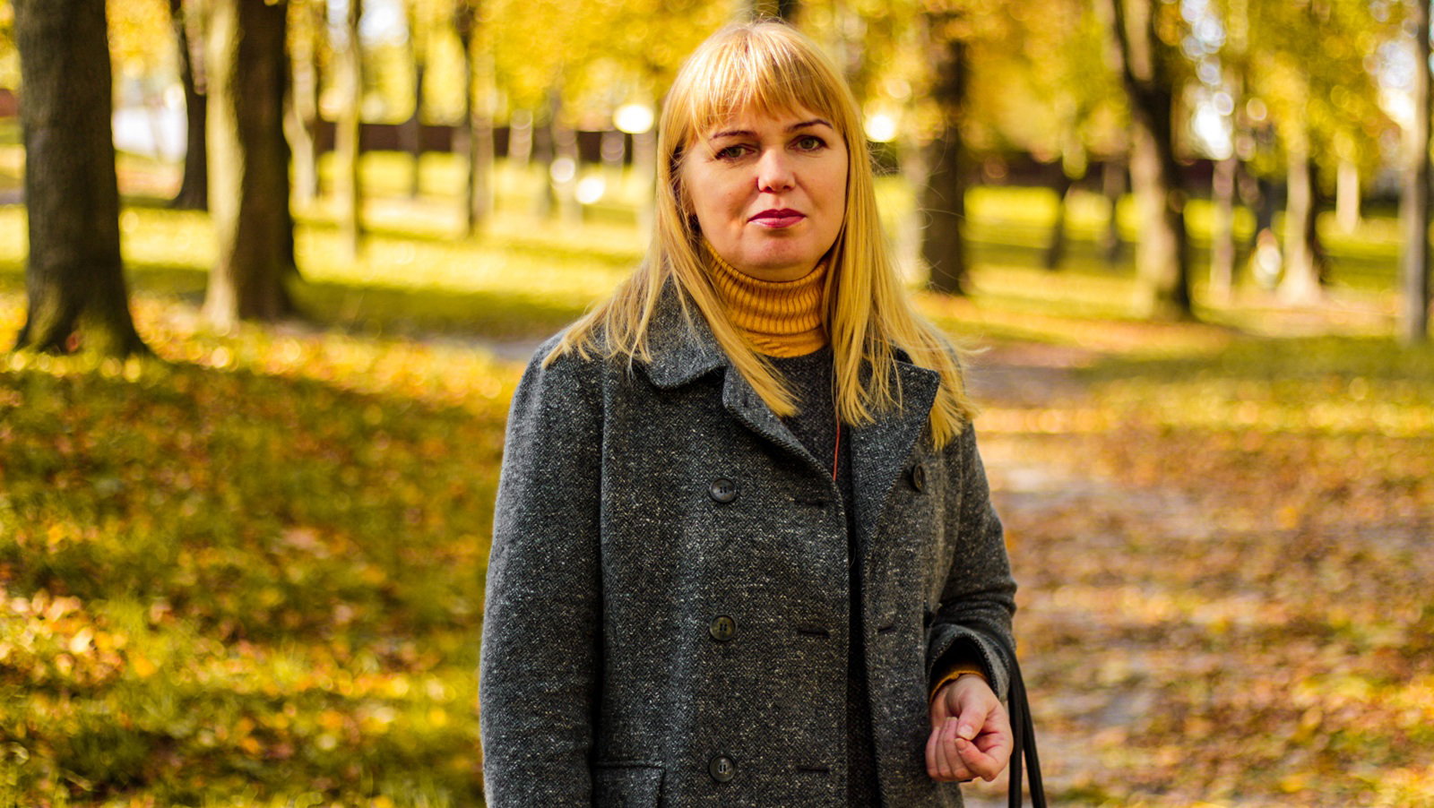 kobieta w  wieku 45 plus blondynka stoi na tle jesiennej alejki z zadowoloną miną i lekkim uśmieszkiem